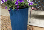 a blue rain barrel with purple flowers and a copper watering can